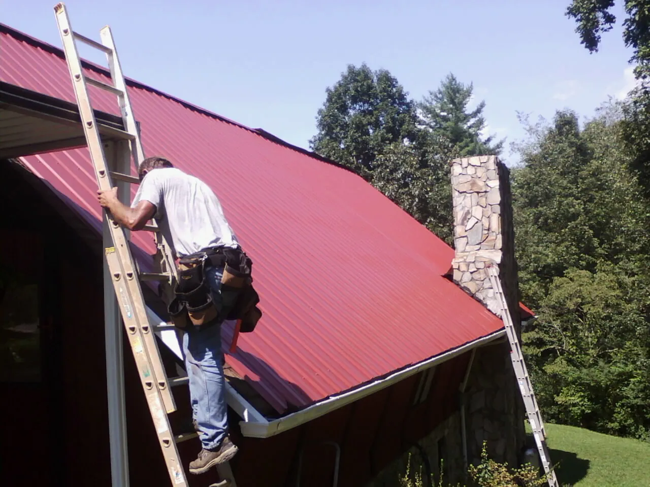Roofer on a ladder beside a red metal roof and stone chimney — Soffit & Fascia Repair by Roofing Brothers in Littlerock